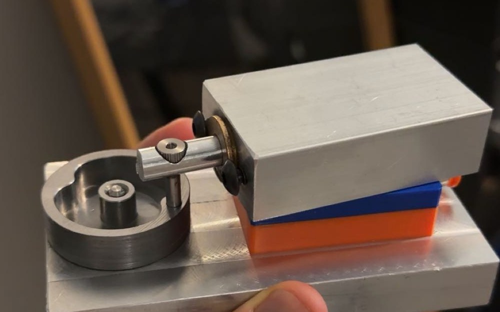 Finished machined compressed-air engine held in hand, showing the aluminum base, flywheel, piston block, and connecting rod